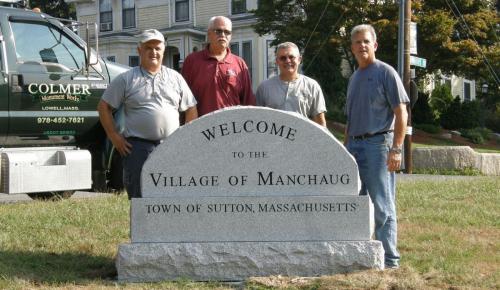 Photo of 4 men facing forward, standing behid granite marker that says &quot;Welcome to the Village of Mantaugh, Town of Sutton MA&quot;