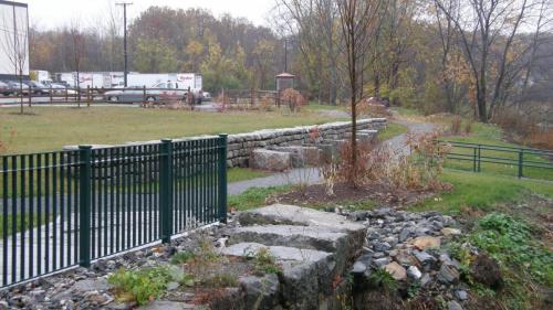 View from inside park toward parking area - - park bounded by stone wall and metal fence in foreground, cars in background